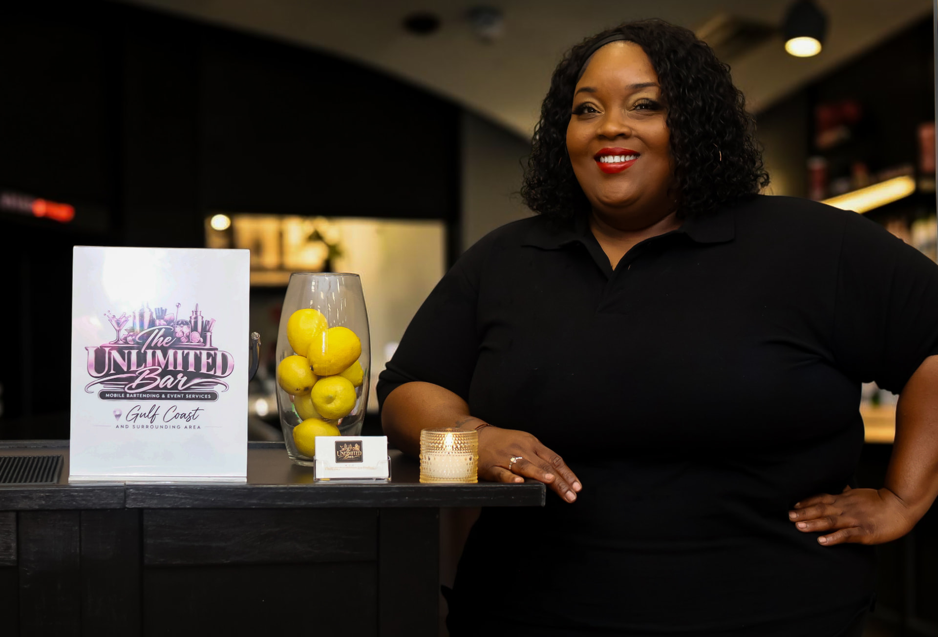 Kaneka Durr behind a styled bar at an event, with The Unlimited Bar tabletop sign and business cards