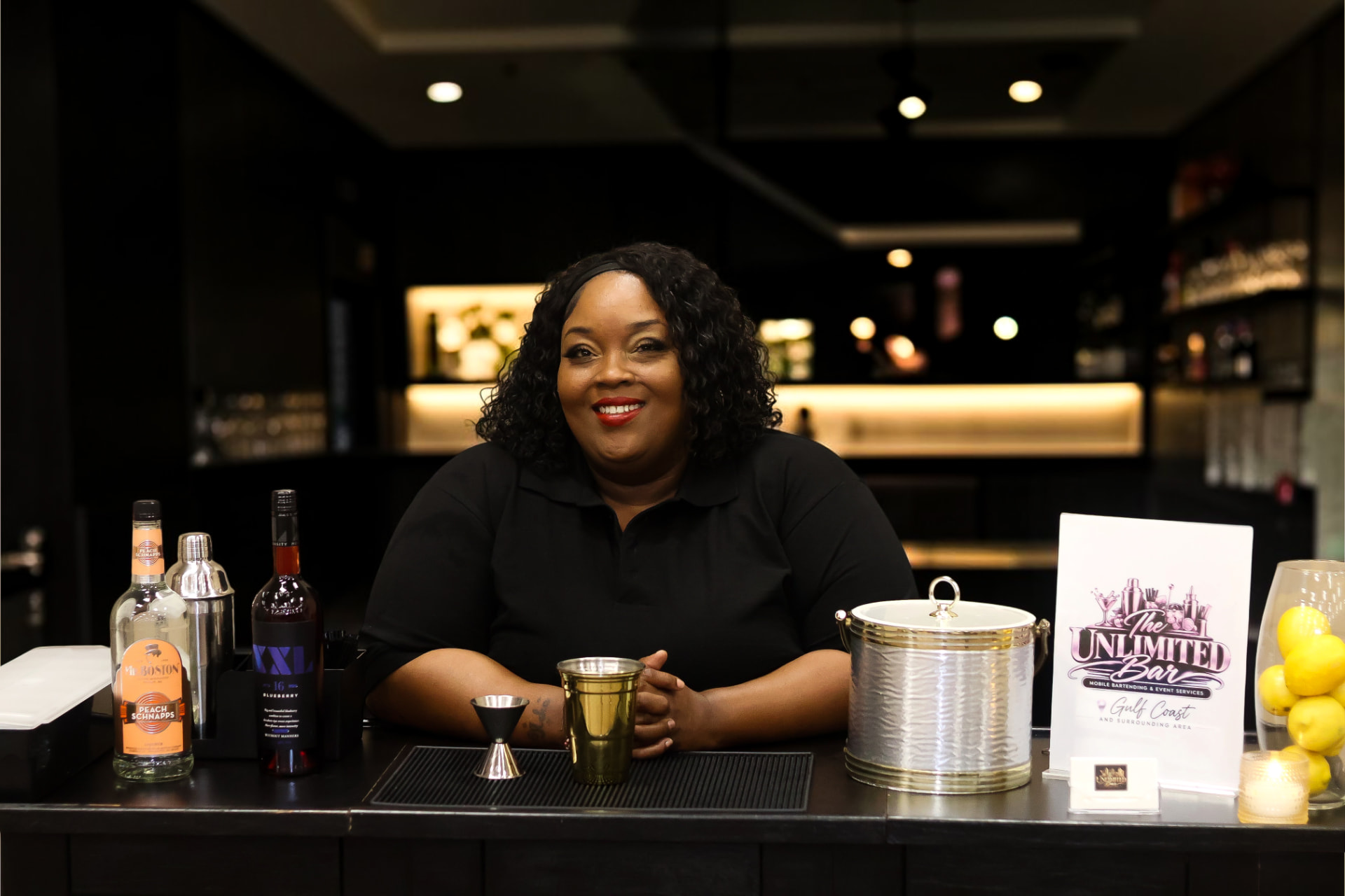 Kaneka Durr behind a stocked bar in a black polo, bottles, shaker and a gold cup on the counter